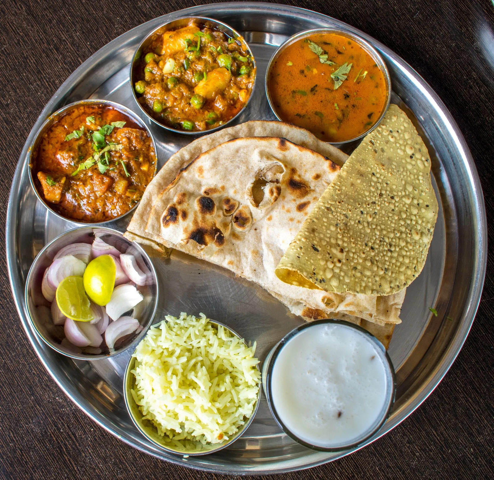 Overhead view of a steel thali with roti, papad, rice, curd, sliced onion and three katoris of curry