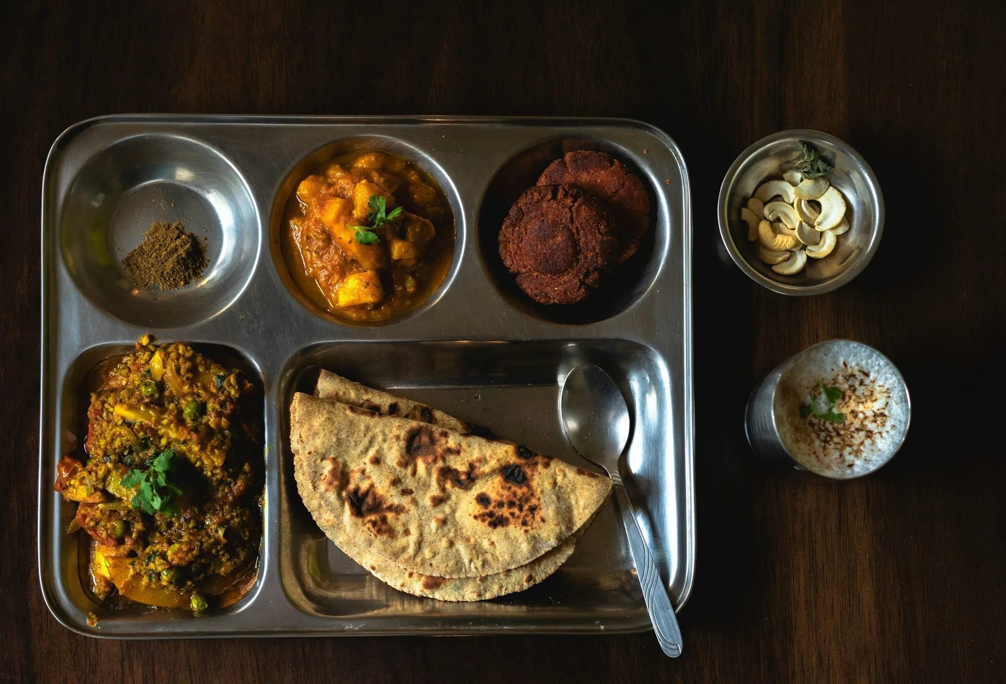 A stainless steel compartment tray with rotis, two vegetable dishes and a glass of buttermilk